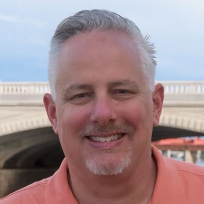 Portrait of Scott Smyth wearing orange shirt in frotn of bridge